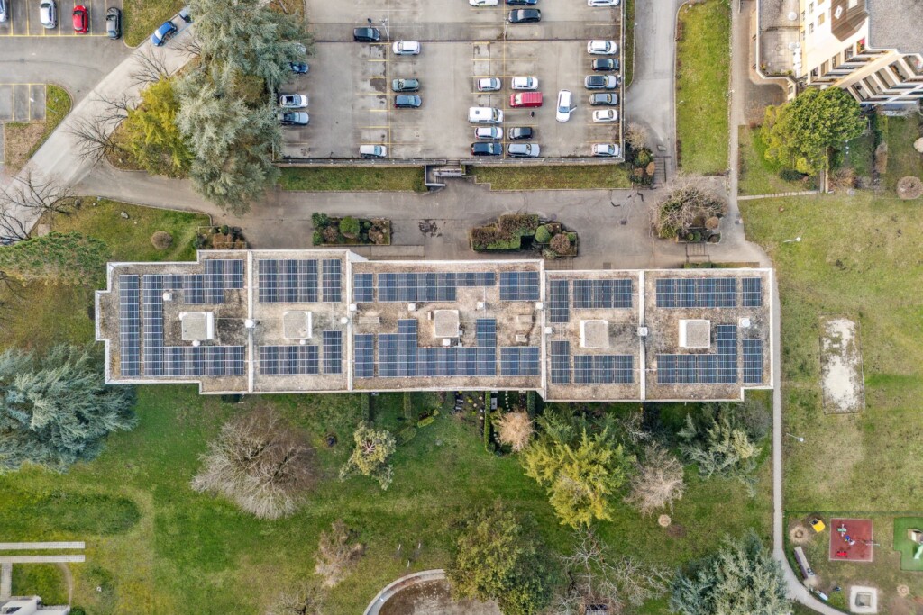 Vue sur le toit de l'immeuble de DSPF à Route de Cité-Ouest 21–29, 1196 Gland. Il y a des panneaux solaires sur le toit et un parking. Parc vert autour.